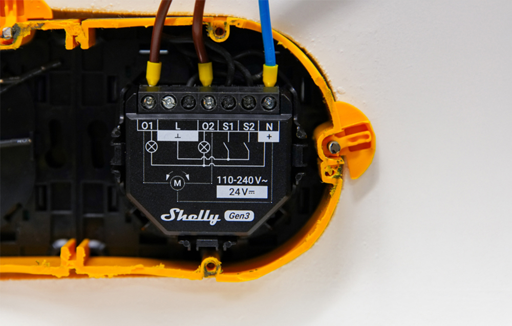 Μονάδα Shelly 2PM Gen3 τοποθετημένη σε ηλεκτρικό κουτί με συνδεδεμένα καλώδια, περιτριγυρισμένη από κίτρινο υλικό τοποθέτησης